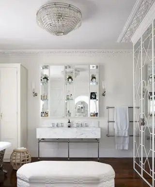 Un baño blanco con lámpara de araña y otomana blanca.