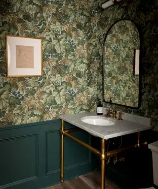 Un ejemplo de decoración de pared de tocador que muestra un tocador de color verde oscuro con papel tapiz botánico verde y un lavabo tradicional.
