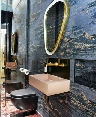 Un tocador oscuro con un inodoro negro, un lavabo rosa y paredes empapeladas de color oscuro.