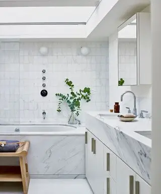 Cuarto de baño elegante con encimera de lavabo de mármol y panel de bañera, azulejos blancos texturizados con una forma geométrica única, dos apliques redondos de ópalo cerca de la ducha y la bañera, jarrón con vegetación junto a la bañera, armario con espejo montado en la pared, asiento de madera junto a la bañera