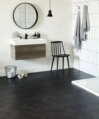 Suelo de vinilo con apariencia de parquet negro en un cuarto de baño con paredes blancas y un lavabo de madera junto a una silla negra