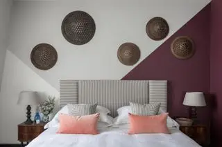 Dormitorio con paredes grises y granate, cojines color melocotón, cabecera a rayas, mesa auxiliar y discos tejidos en la pared.