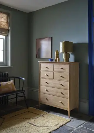 chambre verte avec grand garçon, sol en pierre, tapis, lampe, bases, œuvres d'art, banc et coussin ercol, store à la fenêtre, cadre de porte bleu électrique