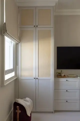 quarto neutro com guarda-roupa embutido, cômoda, TV montada na parede, persianas creme na janela