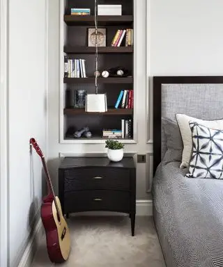 Una idea de dormitorio para adolescentes con una guitarra en el suelo, ropa de cama gris y estantes abiertos de madera oscura integrados en las paredes blancas.
