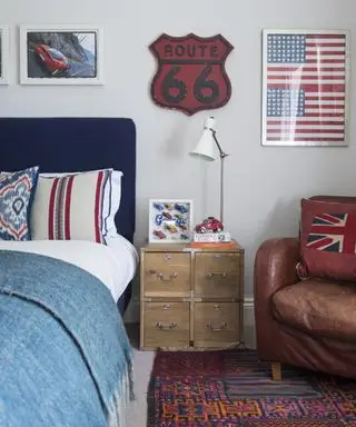 Una idea de dormitorio para adolescentes con cabecera azul, obras de arte americanas, paredes blancas y sillón de cuero marrón.