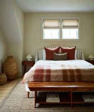 Dormitorio color crema con cama a rayas, manta a cuadros color óxido, almohadas color óxido, mesitas de noche de mediados de siglo y banco de madera.