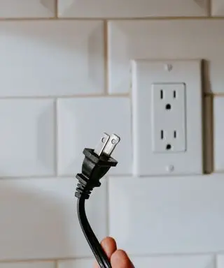 Un enchufe americano negro sostenido frente a un tomacorriente blanco en una pared de azulejos blancos