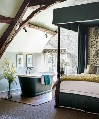 Dormitorio con cama con dosel con tela de William Morris y bañera independiente de color azul oscuro en la casa de campo de los Cotswolds del siglo XII.
