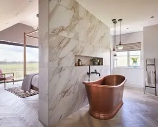 Dormitorio grande y moderno con bañera, separado por una pared divisoria de mármol, bañera de cobre, suelo de madera, trío de luces colgantes, cama y sillón de cuero y madera al fondo, grandes ventanales del suelo al techo.