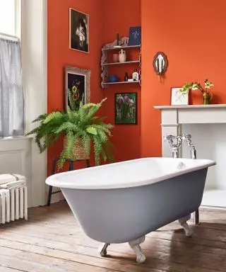 Cuarto de baño pintado de naranja con bañera independiente blanca y gris, suelo de madera oscura, chimenea tradicional, alcoba decorada con cuadros enmarcados, estante de madera con adornos, taburete de madera con planta