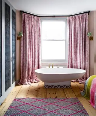 Baño independiente con soporte elevado de azulejos en el ventanal del dormitorio principal, cortinas rosas, dos apliques con pantallas verdes, suelo de madera con alfombra estampada rosa, ropa de cama multicolor