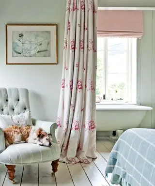 Dormitorio y baño neutros, separados por una cortina floral, bañera con patas, perro en un sillón tapizado, suelos pintados de blanco