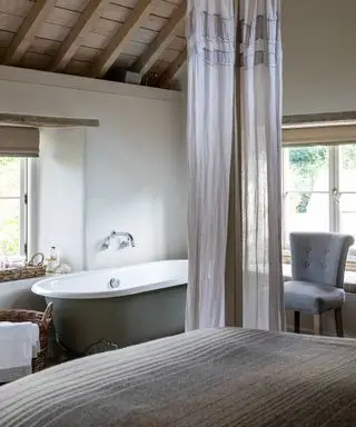 Dormitorio de estilo rural tradicional con cama con dosel y cortinas transparentes, baño pequeño junto a la ventana, sillón junto a otra ventana, techo abovedado de madera