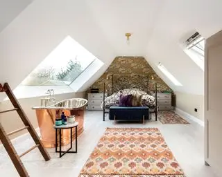 Dormitorio estilo loft con techo abovedado, dos tragaluces, pared empapelada detrás de la cama, somier de metal, dos alfombras estampadas, baño de cobre junto al tragaluz.