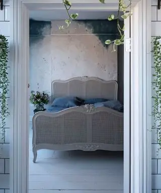 Vista attraverso una porta dal bagno alla camera da letto principale con pavimenti in legno bianco, pareti colorate di blu e letto in canna da zucchero alla francese.