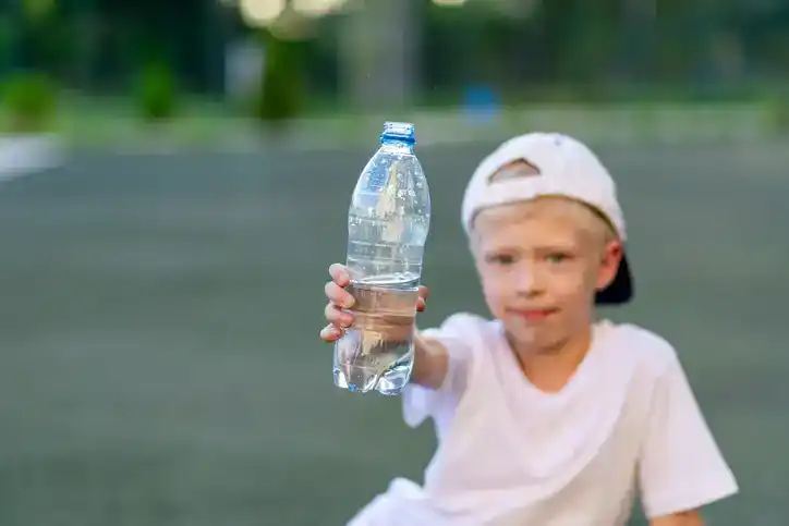 a boy sits on a green lawn on a football field and holds a bottle of water.