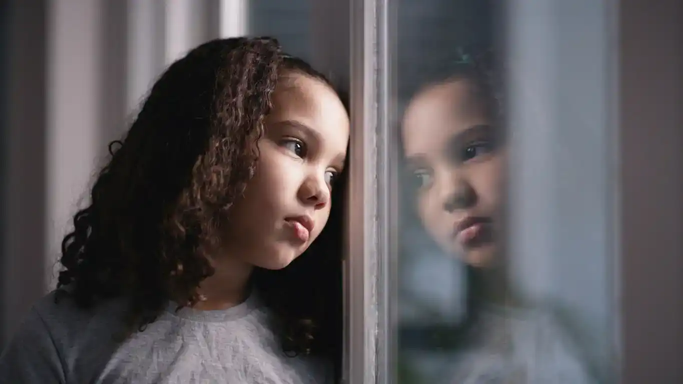 Young girl with curly hair looks sadly out her bedroom window. Concept of generational trauma.