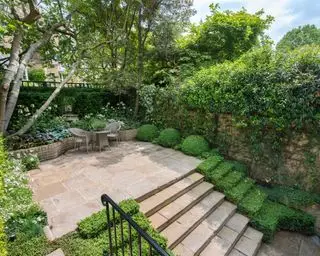 Un ejemplo de cómo diseñar un patio con escalones que conducen a un patio sombreado con sillas, una mesa pequeña y macizos de flores profundos.
