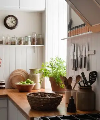 Una cocina de estilo rústico, con plantas en el alféizar de la ventana, cuchillos colgados en la pared y una cesta tejida sobre la encimera.