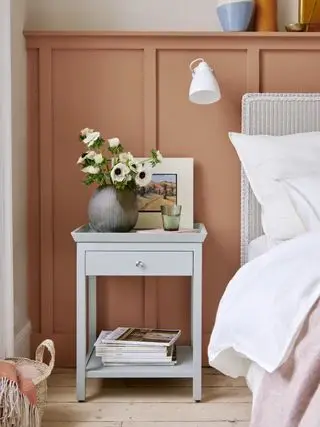 paneles de pared pintados de terracota en el dormitorio, mesita de noche gris pálido, lámpara de pared blanca, cama de mimbre pintada de blanco, ropa de cama blanca, manta rosa, estante encima del panel, piso de madera