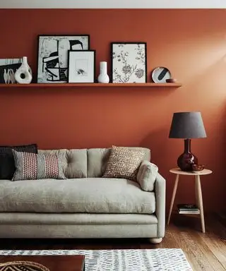 En beige sofa foran en rød vegg med en enkelt svart hylle som viser kunstverk