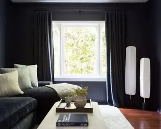 Gemütliches Wohnzimmer mit schwarz gestrichenen Wänden, schwarzen Vorhängen, schwarzem Sofa, dekoriert mit cremefarbenen Kissen und Überwürfen, dunklem Holzboden, grauem Teppich, cremefarbenem Polsterhocker und zwei weißen Laternen-Stehlampen