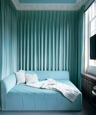 Gästezimmer mit helltürkisfarbenem, quadratischem Samtsofa mit diagonalen Nähten und plissiertem Vorhang an den Wänden