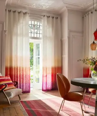 Großer traditioneller Speisesaal mit farbenfrohem, modernem Design. Vorhänge in Ombre-Rosa, Orange und Weiß, heller Holzboden, rosa gestreifter Teppich, runder Esstisch aus Metall mit Samtkäfer-Esszimmerstühlen