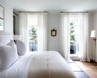 Doppelbett in einem hellgrauen Schlafzimmer mit Holzboden, cremefarbener und weißer Bettwäsche und weißen Vorhängen, die vom Boden bis zur Decke reichende Fenster verkleiden.