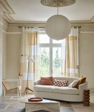 Beruhigendes, neutrales, modernes Wohnzimmer mit Vorhängen mit abstraktem Quadratmuster, hellem Holzboden, abgerundetem Papierlaternenschirm, geschwungenem cremefarbenem Sofa, Stehlampe, Ledersessel, Marmor-Couchtisch auf Teppich