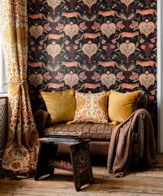 Maximalistisches Wohnzimmerdesign, gemusterte Tapete im William-Morris-Stil, orange und gelb gemusterte Vorhänge, kleines gemütliches braunes Ledersofa mit gelben Kissen und braunem Überwurf, hölzerner Beistelltisch mit aufgemalten Blumendetails