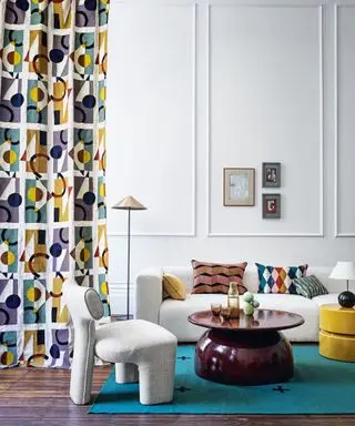 Modernes Wohnzimmer mit weiß gestrichenen Wänden und Paneelen, blauem und gelbem Vorhang mit geometrischem Muster, dunklem Holzboden, blauem Teppich, cremefarbenem, klobigem Sofa und Sessel, gelber Ottomane, Tischlampe und Stehlampe mit Kegelschirm, dunkelbraun glänzendem, abgerundetem Couchtisch