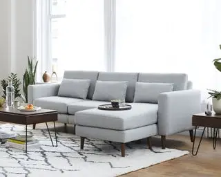 Sofá modular cinza em sala moderna, tapete creme, piso de madeira escura, plantas e mesinhas laterais