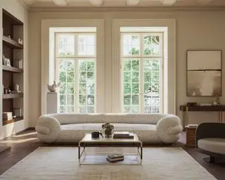 Grande sala de estar neutra com sofá curvilíneo creme, duas janelas grandes, grande tapete creme, mesa de centro de metal