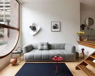 Sofá grande acolchoado cinza na sala de estar rebaixada, piso de madeira, tapete azul, mesa lateral vermelha, grandes janelas em arco, obras de arte nas paredes
