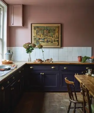 Cozinha pintada de rosa com armários azuis, bancada de madeira, mesa de jantar de madeira, obras de arte na parede