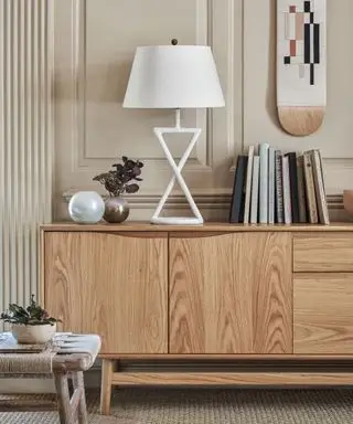 Parede com painéis off white, aparador com acabamento em madeira e luminária geométrica branca, vaso redondo de metal com folhas roxas secas, bola de vidro branca e arte moderna na parede.