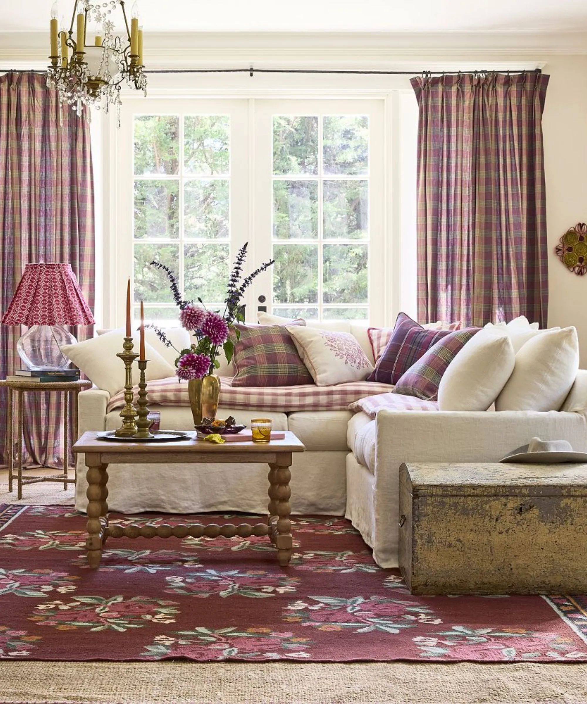 Neutrales Wohnzimmer mit großen französischen Türen und einer weißen Eckcouch mit rosa karierten Kissen und passenden Vorhängen, Holzmöbeln im Vintage-Stil und einem floralen Flachgewebeteppich' loading='lazy' title=