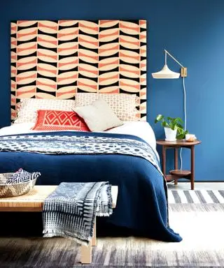 : Un dormitorio con una cabecera alta, paredes pintadas de azul medio y colchas y ropa de cama estampadas. Una cesta y un banco.