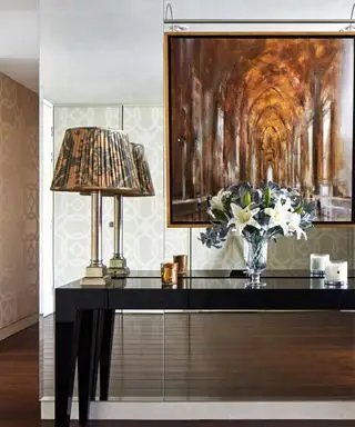 Pared de espejo detrás de un aparador lacado en negro con lámparas que flanquean una pintura en perspectiva de tono marrón y un piso de madera oscura