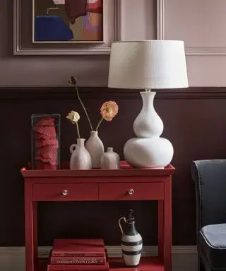 Lys lilla malt rom med mørk lilla malt panel, malt bilde på vegg, hvit lampeskjerm, rødt sidebord med bøker og dekorative ornamenter, blå stol