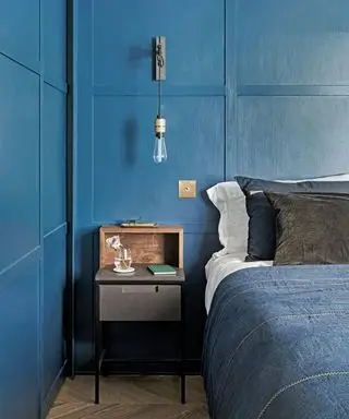 Soverom med blå panelvegger, dobbeltseng med blå denim sengetøy og blå og sorte puter, vegglampe med synlig hengende lyspære, nattbord i tre og tregulv