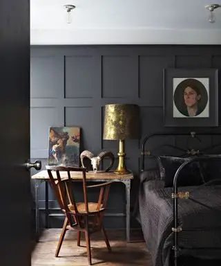 Soverom med svartmalt panel, sengeramme i sort metall med sort dyne, skrivebord i tre og metall med tradisjonell lenestol i mørkt tre, malerier på skrivebord og på vegger, metallisk lampeskjerm