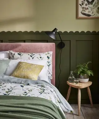 Soverom med mørk grønn veggpanel med kamskjell, lysegrønnmalte vegger, svart vegglampe, lys trekrakk, grønt og grått blomstersengetøy med blekrosa hodegjerde, spredeputer