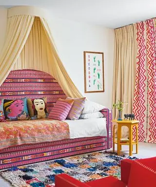 Høj pink mønstret daybed med creme baldakin, dekoreret med farverige puder og plaider, farverigt ternet tæppe, creme tæppe, rød lænestol, pink mønstrede og creme gardiner