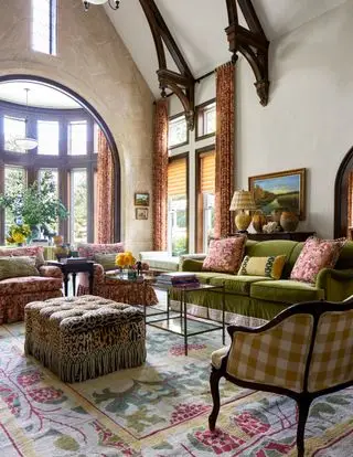sala de estar com sofá verde, poltronas florais, poltrona xadrez, pufe com estampa de leopardo, mesa de centro, tapete colorido, teto abobadado, obras de arte, cortinas e persianas