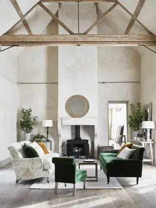 sala neutra com vigas, lareira, sofá e poltrona verdes, sofá com estampa floral verde, tapete, plantas, consoles, mesa lateral, tapete, piso de madeira, espelho