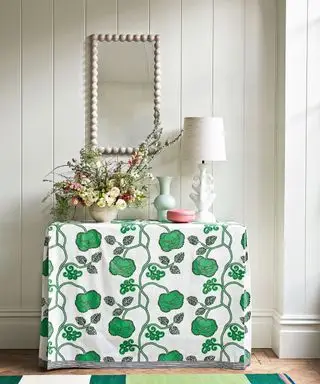 mesa de console no corredor com toalha de mesa floral verde e branca, espelho emoldurado em bobina na parede branca, abajur creme, vaso de flores