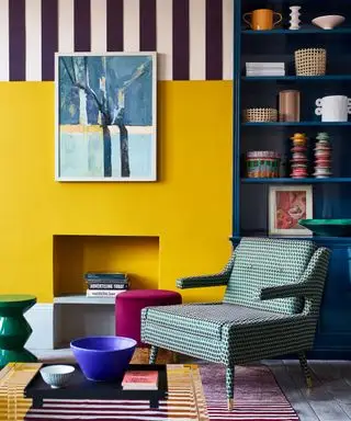 Farverigt hjørne af en stue, gul malet væg, pejs, polstret lænestol, stiftet malet design på overvæggen, blåmalet reol, kunst på vægge, bøger og ornamenter på hylde